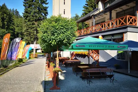 Taras restauracyjny z ławkami i stołami, parasolami, obok łuki startowe i flagi.