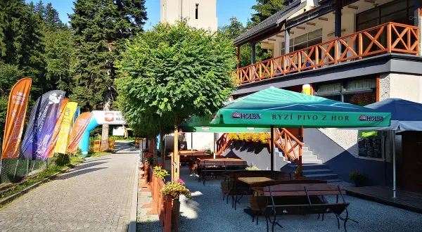 Taras restauracyjny z ławkami i stołami, parasolami, obok łuki startowe i flagi.