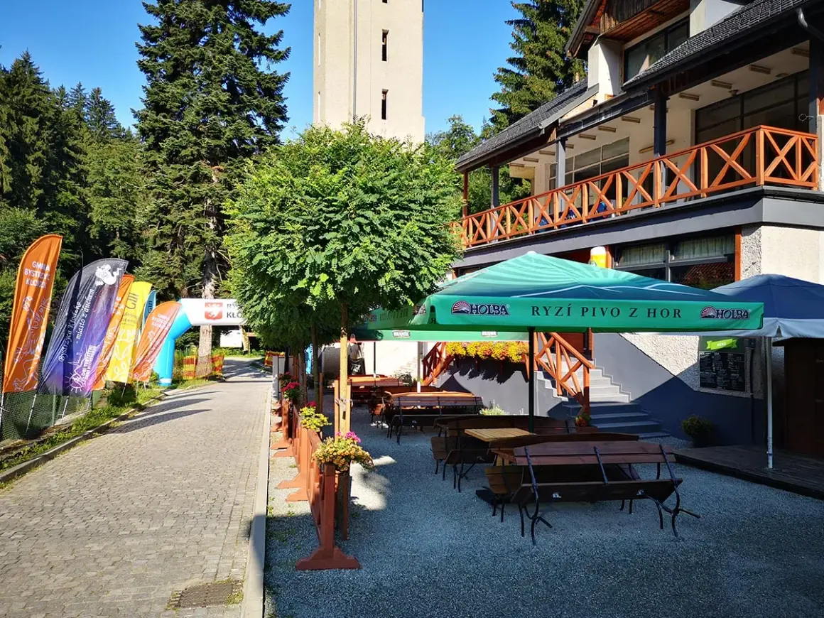 Taras restauracyjny z ławkami i stołami, parasolami, obok łuki startowe i flagi.