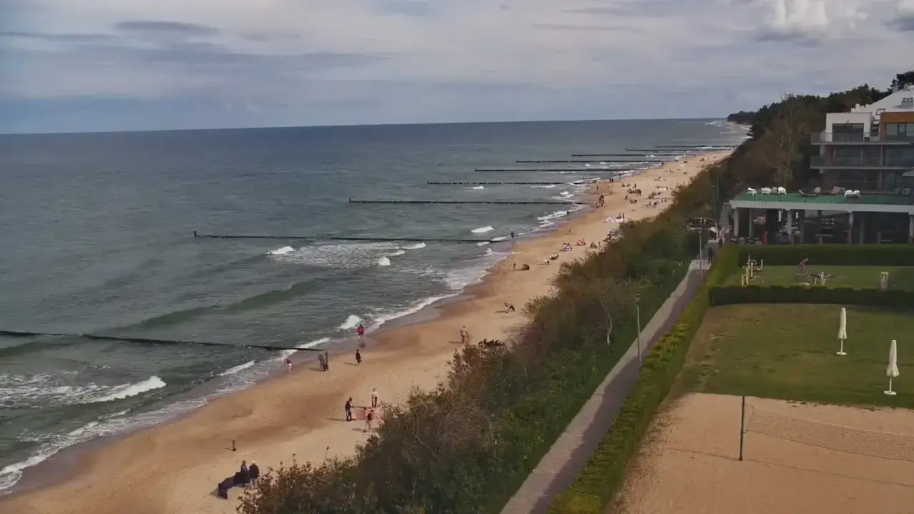 Plaża w Ustroniu Morskim tętni życiem. Spacerowicze i plażowicze korzystają z uroków Bałtyku, a w tle widać hotel i tereny rekreacyjne.